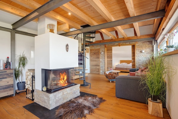 A modern living room with a fireplace, wooden beams, a stone wall, and a staircase leading to another level.