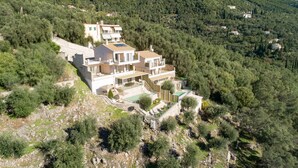 Une maison à plusieurs étages avec une piscine, entourée d’une forêt dense et de collines.