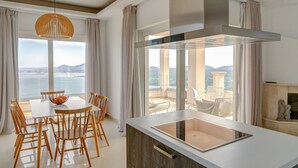 Un espace repas moderne avec une table et des chaises, un saladier rempli de fruits et une vue sur la mer grâce à de grandes fenêtres.