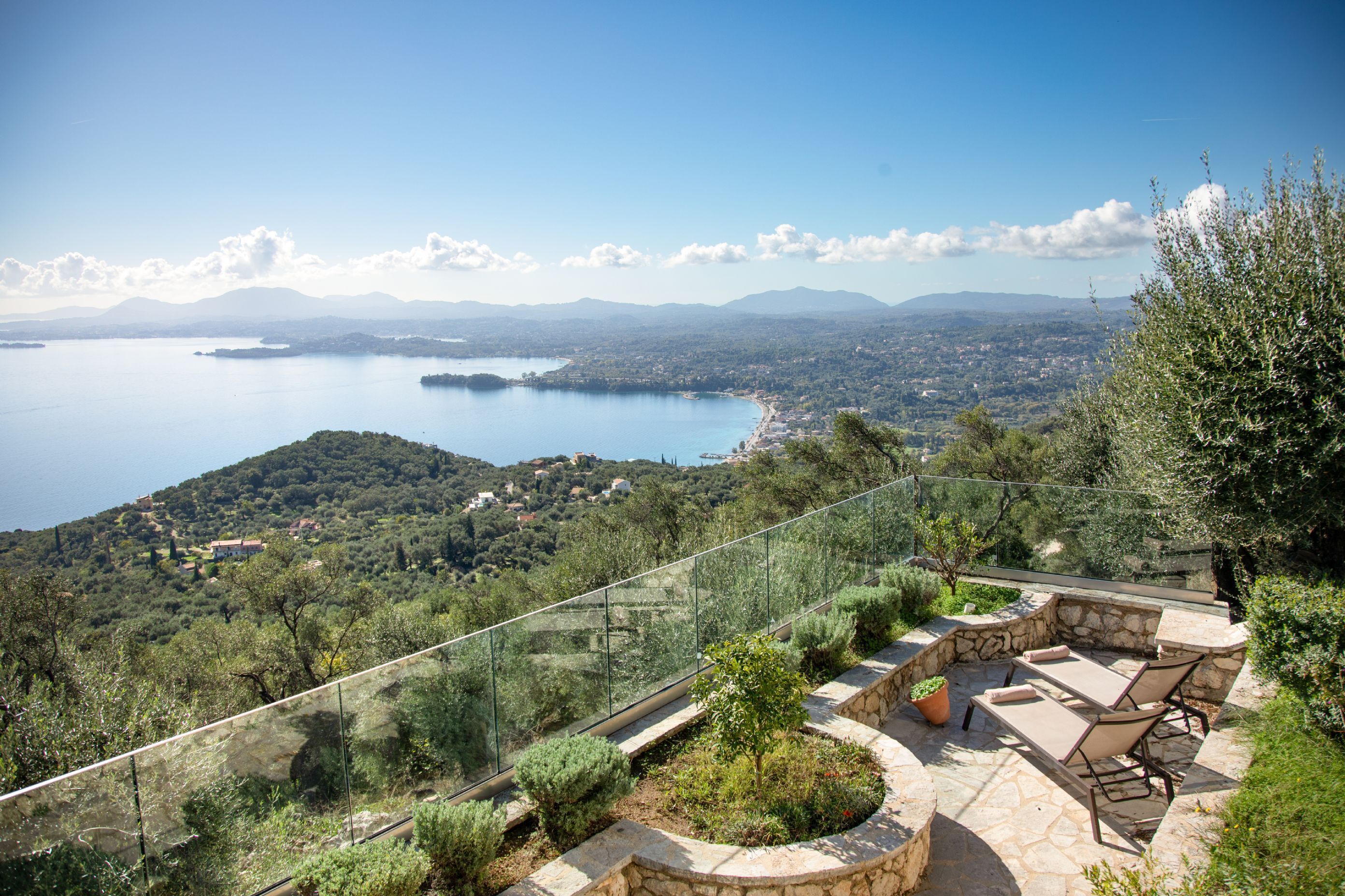 A patio with stone flooring and a glass railing overlooking a scenic view of a lake and distant mountains.