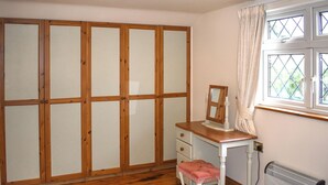 Ein Schlafzimmer mit Holzschrank, Nachttisch mit Spiegel, Heizkörper und einem Fenster mit Vorhängen.