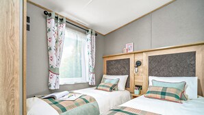 Ein Zimmer mit zwei Betten, einem Fenster mit blumigen Vorhängen, einem Kopfteil mit einer Leuchte und einem kleinen Tisch mit Getränkehalter.