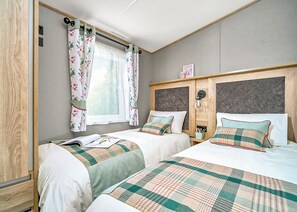 Ein Zimmer mit zwei Betten, einem Fenster mit blumigen Vorhängen, einem Kopfteil mit einer Leuchte und einem kleinen Tisch mit Getränkehalter.