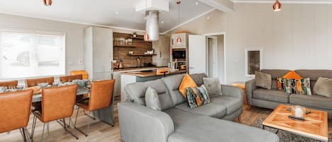 A modern living room with a grey sofa, dining table, and chairs, a kitchen area, and pendant lighting.