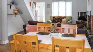 Un coin repas avec une table et des chaises en bois, un escalier, un salon avec un canapé et un téléviseur, et une fenêtre avec des stores.