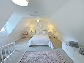 Un dormitorio con techo inclinado, una cama, dos mesitas de noche con lámparas, una silla, un tapete y un banco empotrado con toallas.