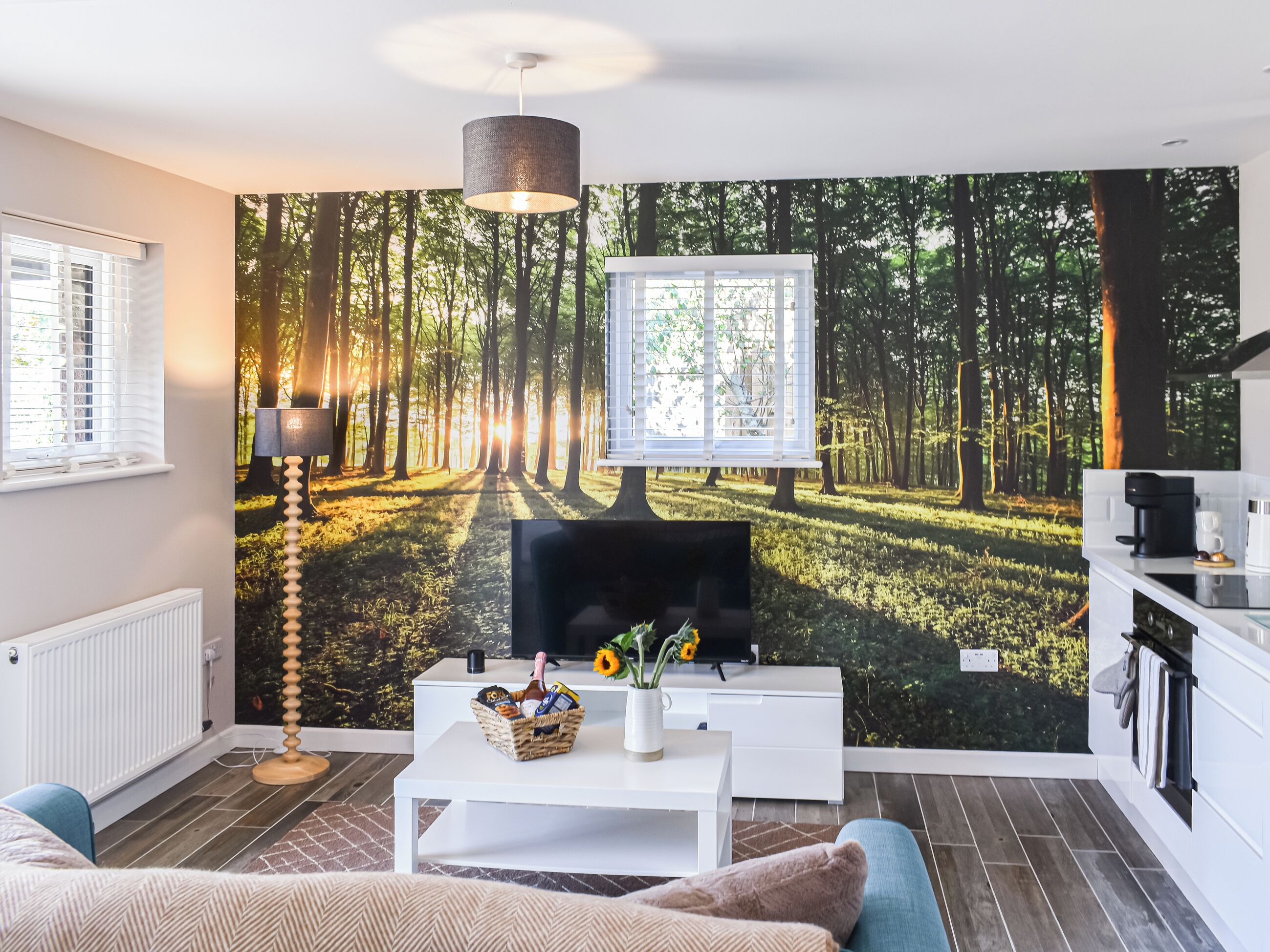 A modern living room with a forest-themed wallpaper, a white sofa, a coffee table with a basket, a floor lamp, and a kitchen area with a coffee machine.