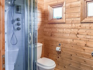 Une salle de bain rustique, avec une cabine en bois, une douche fermée par des parois de verre, des toilettes et deux petites fenêtres.