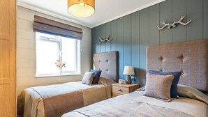 Een slaapkamer met twee bedden, een raam met zonwering, een lamp op een nachtkastje, en decoratieve geweihandelingen aan de muur.