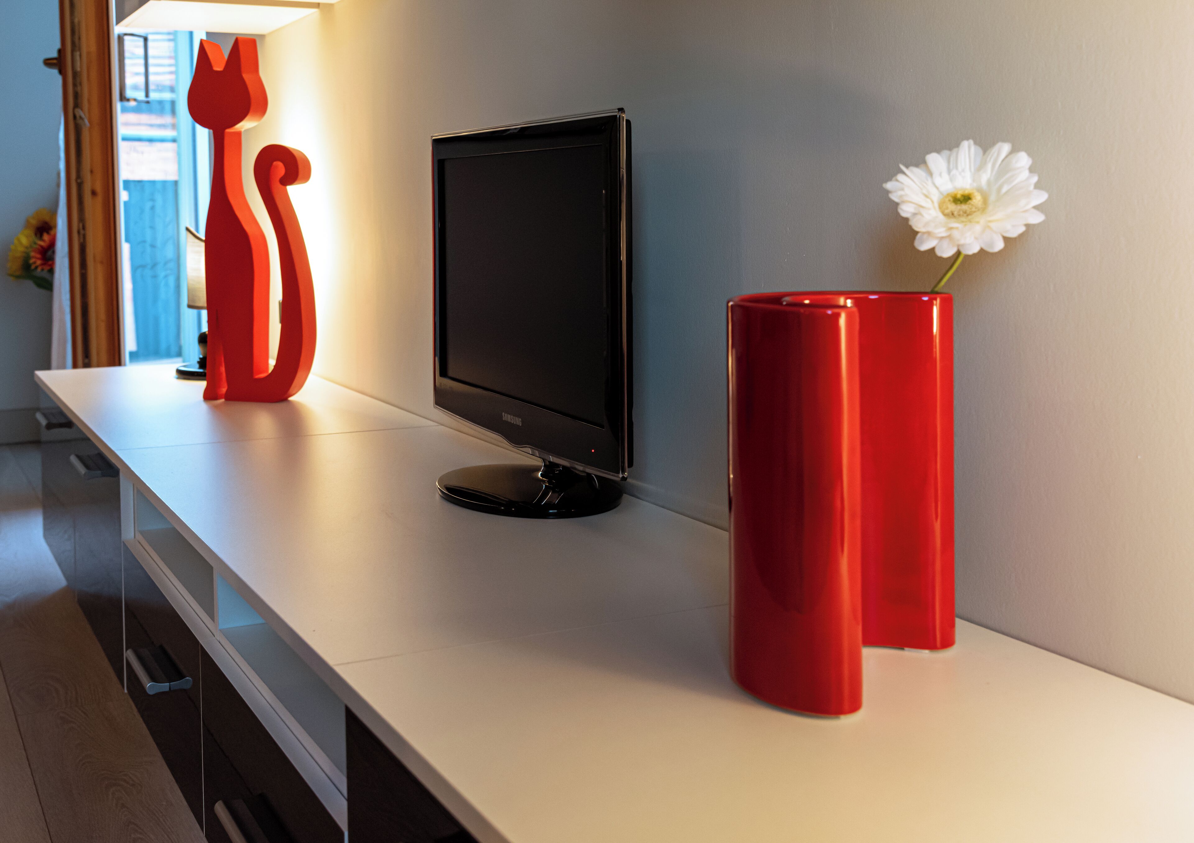 A modern living room with a flat-screen TV, a red vase with a white flower, and a red cat-shaped light on a white table.