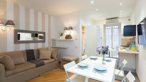 A modern living room with a sofa, a dining table set for four, and a kitchen area with a TV and appliances. - Rental in Rome Vittoria Apartment (Rome)
