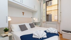 Un letto rifatto con due asciugamani, una lampada da comodino e una piccola pianta su un tavolino.