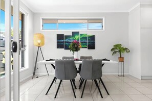 A modern dining room with a white table, four grey chairs, a floor lamp, and a potted plant. - Reef Retreat Modern Beach HouseSanctuary (Port Noarlunga)