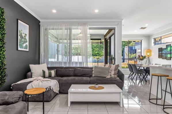 A modern living room with a grey sectional sofa, a white coffee table, and a dining area with bar stools. - Reef Retreat Modern Beach HouseSanctuary (Port Noarlunga)