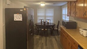 Eine kleine Küche mit Kühlschrank, Esstisch mit Stühlen, Holzschränken und einer Mikrowelle.