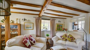 A cozy living room with two white sofas, a small wooden table, and a wicker stool. There are wooden beams on the ceiling, a bookshelf, and a window with curtains. - 2 Bed in Stiffkey (oc-1892) (Stiffkey)