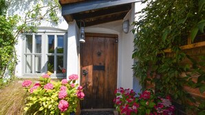 A rustic wooden door with a metal knocker, a small porch area with a doormat, and potted plants with pink flowers. - 2 Bed in Stiffkey (oc-1892) (Stiffkey)