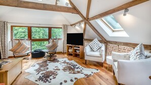 A loft-style room with exposed wooden beams, a large window with a view of trees, a flat-screen TV, and two chairs with patterned cushions.