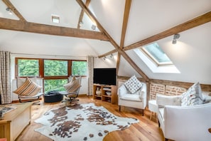 A loft-style room with exposed wooden beams, a large window with a view of trees, a flat-screen TV, and two chairs with patterned cushions. - 3 Bed in Ketteringham (oc-1878) (Ketteringham)