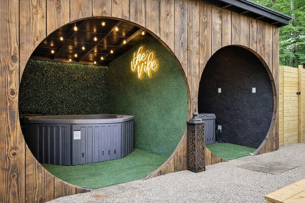 A wooden structure with two circular openings, one with a hot tub and a neon sign, and the other with a lantern.