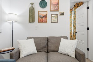 A living room with a sofa, two pillows, a lamp, and wall decorations including framed pictures and a macrame wall hanging.