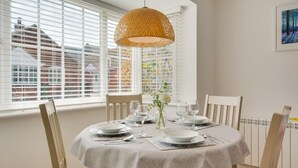 Un coin repas avec une table ronde dressée pour quatre personnes, des chaises blanches, un luminaire suspendu en fibres tressées et une vue sur les bâtiments à travers de grandes fenêtres équipées de stores.