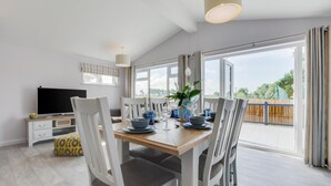 A dining area with a wooden table, white chairs, and a TV on a white cabinet. - 3 Bed in St. Merryn (oc-p00701) (Saint Merryn)