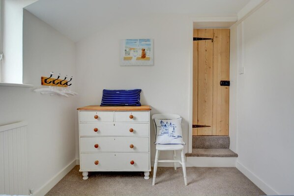 A small, white room with a wooden dresser, a chair, and a door leading to another room.