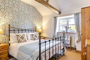 A bedroom with a patterned wallpaper, a bed with a headboard, a window with curtains, and a wooden wardrobe. - 2 Bed in Swanton Abbott (oc-1173) (Swanton Abbott)