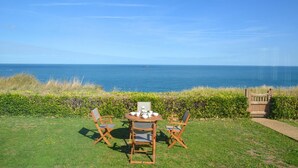 A grassy outdoor area with a wooden table and chairs, a bench, and a view of the ocean. - 3 Bed in Cromer (oc-1909) (Cromer)