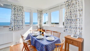 A dining area with a table set for four, large windows with curtains, a wooden chair, and a small desk with a telephone. - 3 Bed in Cromer (oc-1909) (Cromer)