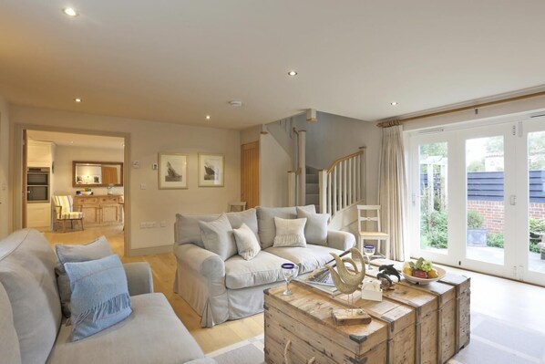 A spacious living room with a grey sofa set, a wooden coffee table, and a staircase leading to a balcony. - 2 Bed in Thorpeness (oc-vels) (Thorpeness)