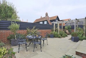 A patio with a round table and chairs, surrounded by potted plants and a brick wall. - 2 Bed in Thorpeness (oc-vels) (Thorpeness)