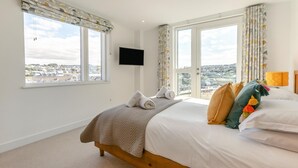 A hotel room with a bed, a television, and a view of a cityscape through large windows. - 3 Bed in Perranporth (oc-p00695) (Perranporth)