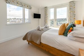 A hotel room with a bed, a television, and a view of a cityscape through large windows. - 3 Bed in Perranporth (oc-p00695) (Perranporth)
