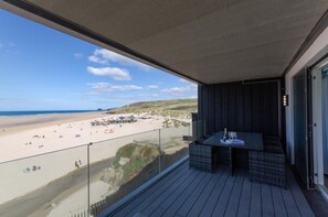 A balcony with a view of a beach and ocean, a table with a bottle and glasses, and a glass railing.