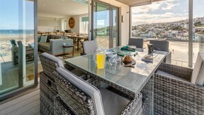 A sunlit terrace with a glass table, wicker chairs, and a view of the beach and town. - 3 Bed in Perranporth (oc-p00695) (Perranporth)