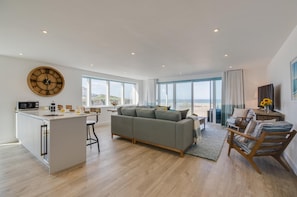 A spacious living area with a large window, a grey sofa, a wooden coffee table, and a kitchen area with a microwave.