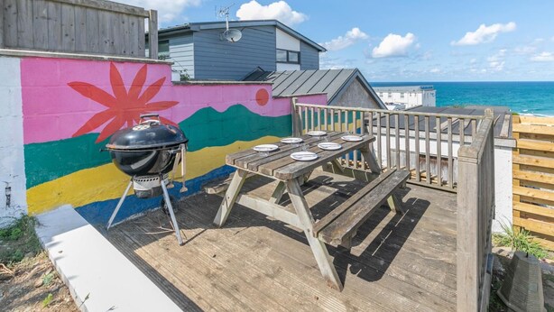Eine Holzterrasse mit Grill und Esstisch, mit Blick auf ein farbenfrohes Wandgemälde und das Meer.