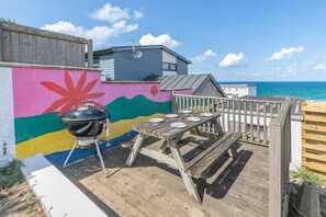 A wooden deck with a barbecue grill and a table set for dining, overlooking a colorful mural and the sea. - 5 Bed in Newquay (oc-p00894) (Newquay)