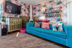 A living room with a teal sofa, colorful pillows, a flat-screen TV, a white cabinet, and a patterned wall. - 5 Bed in Newquay (oc-p00894) (Newquay)