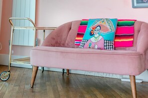 Een roze bank met een kleurrijk kussen en een metalen karretje in een kamer met houten vloeren.