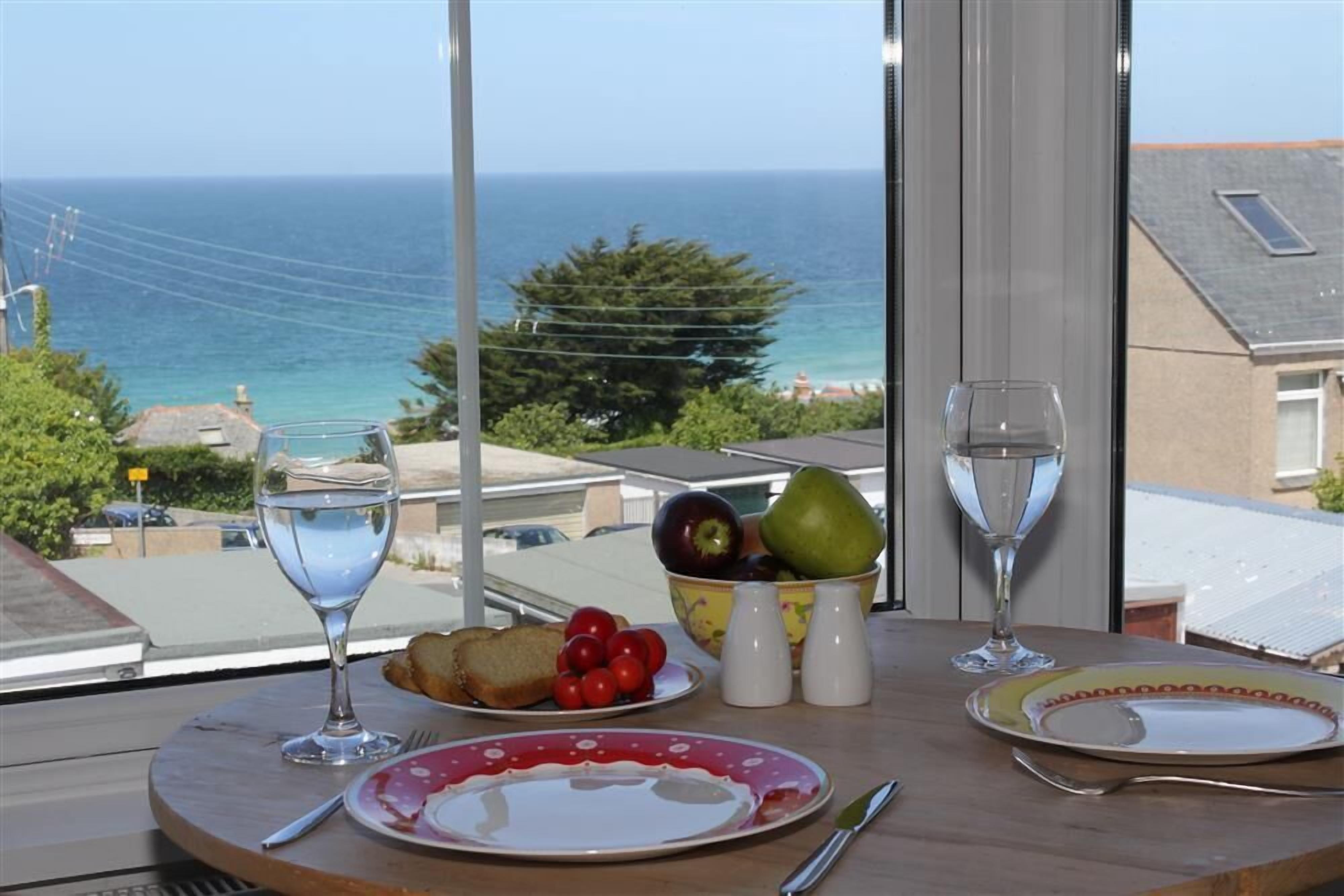A table with a view of the sea, a bowl of fruit, two glasses of water, and a plate with toast and tomatoes.