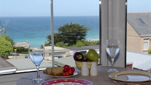 Una mesa con vista al mar, un cuenco de fruta, dos vasos de agua y un plato con tostadas y tomates.