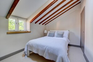 A bedroom with a sloped ceiling, a bed with white bedding, a window with blinds, and a wooden beam structure. - 3 Bed in North Creake (oc-1916) (North Creake)