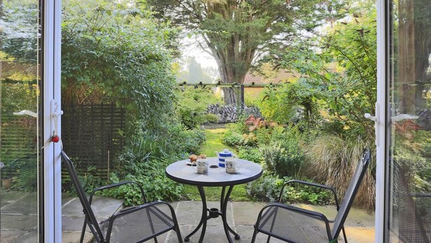 Een terras met een tafel en twee stoelen, omringd door weelderige begroeiing en een grote boom.