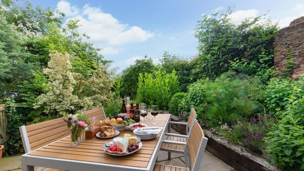 A wooden table set outdoors with a variety of fruits, cheeses, and bottles, surrounded by lush greenery and a brick wall. - 3 Bed in Crayke (oc-y056) (Crayke)