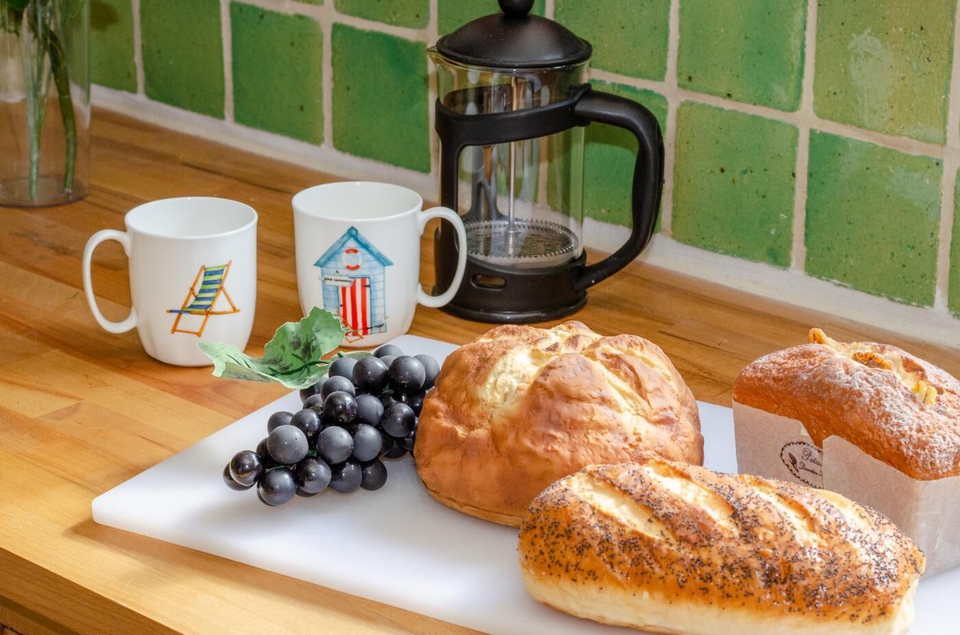 A breakfast setting with a coffee maker, two mugs, a loaf of bread, a pastry, and a bunch of grapes on a wooden table.