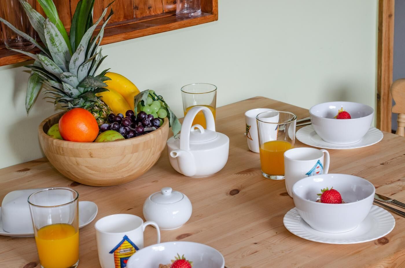 A breakfast setting with a wooden table, a fruit bowl, white cups, a teapot, and a glass of orange juice.