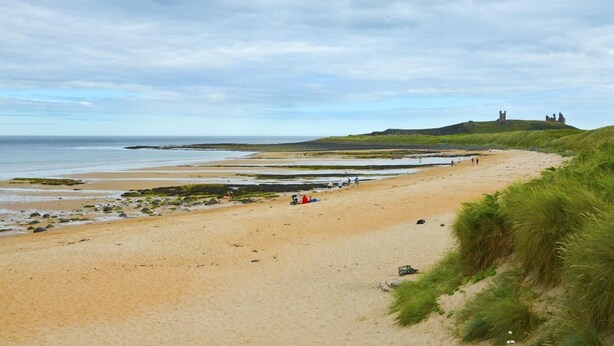A sandy beach with grassy dunes, a few people, and distant ruins on a hill. - 1 Bed in Alnwick (oc-nb312) (Embleton)